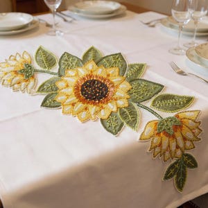 May include: A white table runner with a beaded sunflower design. The sunflowers are yellow, orange, and brown with green leaves. The runner is placed on a white tablecloth, with plates, glasses, and silverware visible.
