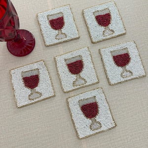 Puede incluir: Seis posavasos cuadrados con cuentas con un diseño de copa de vino. Los posavasos tienen un fondo blanco, un diseño de copa de vino tinto y un borde dorado. Una copa de vino tinto es visible en la esquina superior izquierda.