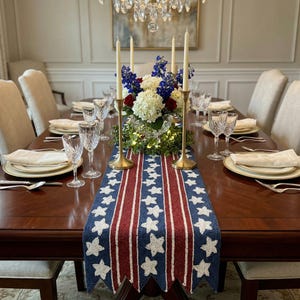 May include: A formal dining table set for a special occasion. The table features a patriotic-themed runner with red and blue stripes and white stars. Tall gold candlesticks hold white candles, and a floral arrangement sits in the center.
