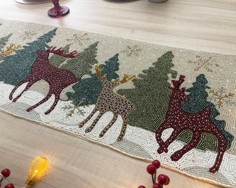 May include: Beaded Christmas table runner featuring reindeer and evergreen trees in a winter scene. The runner has a beige background with white, green, brown, and burgundy beads. Snowflakes and a snowy ground complete the festive design.