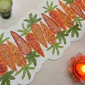 Puede incluir: Camino de mesa con cuentas con un diseño de zanahorias en tonos naranja, rojo y amarillo, con hojas verdes. El camino de mesa tiene un borde festoneado y está sobre un fondo neutro. Ideal para decorar una mesa.