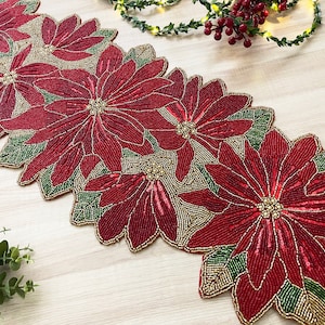 Op de afbeelding: Een rode en gouden kralen tafelrunner met een kerstster ontwerp. De runner is gemaakt van kleine kralen die aan elkaar zijn genaaid om een ​​gedetailleerd bloemenpatroon te creëren.