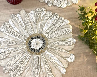 May include: A white beaded table mat with a large flower design. The center of the flower is a circle of gold, black, and white beads.