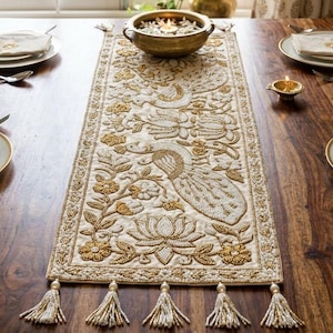 Könnte beinhalten: Ein Holztisch, der für eine Mahlzeit gedeckt ist, mit einem cremefarbenen Tischläufer mit goldener Pfau- und Blumenstickerei. Der Tisch ist mit Tellern, Servietten, Besteck und einer dekorativen Schale gedeckt. Auf dem Tisch brennt eine Kerze.