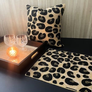 Puede incluir: Una almohada de tiro de leopardo con cuentas negras y doradas y un mantel individual a juego. La almohada está sobre una bandeja de madera con dos candelabros de vidrio transparente y una vela dorada.