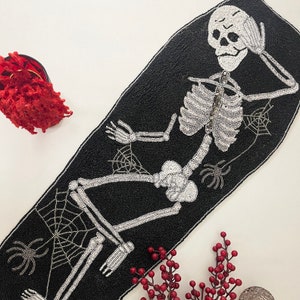 Peut inclure: Un chemin de table noir et blanc en perles avec un motif de squelette. Le squelette est allongé, les bras croisés et la tête reposant sur sa main. Le chemin de table est décoré de toiles d'araignées et de perles.