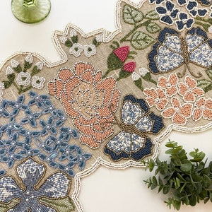 Op de afbeelding: Een geperst tafelloper met een bloemenmotief met vlinders, rozen en bladeren in tinten blauw, roze en groen.