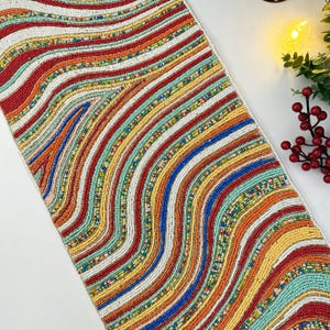 May include: A beaded table runner with a vibrant, wavy striped design. The runner showcases a mix of colours, including red, orange, yellow, blue, and green. The beaded texture adds visual interest. Ideal for adding a colourful touch to a dining table, it measures approximately 150 cm in length.