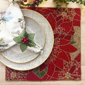 May include: A red and gold beaded placemat with a poinsettia design. A white napkin with a floral pattern is folded on a white and gold plate.