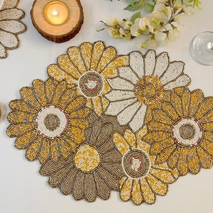 May include: A set of four beaded placemats featuring a sunflower design in shades of yellow, white, and brown. The placemats are made of small beads sewn together to create a textured, floral pattern.
