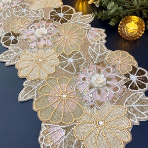 Puede incluir: Camino de mesa floral con cuentas doradas, beige y rosas. El camino presenta intrincados diseños de flores y hojas, con una apariencia texturizada y dimensional. Las flores están adornadas con cuentas y lentejuelas, creando un acento decorativo.