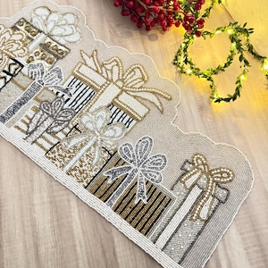 May include: Beaded Christmas table runner featuring gift box designs in gold, silver, and white. The gifts have ribbon bows and are arranged on a neutral background. The runner is edged with a beaded border.