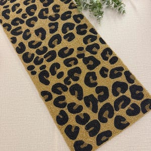 May include: A beaded table runner with a leopard print design. The runner features a tan background with black spots. The beads are sewn together to create the pattern. The runner is rectangular and appears to be made of fabric.