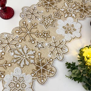 May include: A beige table runner with a scalloped edge is decorated with white and gold beaded snowflakes and stars. The runner is perfect for adding a festive touch to your holiday table.