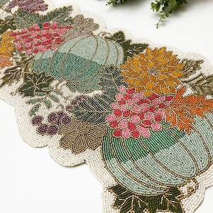 May include: Beaded table runner with a fall theme. The runner features pumpkins, leaves, and flowers in shades of green, gold, pink, and burgundy.