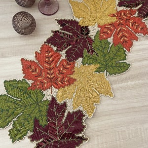 Pode incluir: Um caminho de mesa com contas com desenhos de folhas de bordo em cores de outono. As folhas são em tons de verde, dourado, laranja e bordô, com intrincados trabalhos de contas e uma borda branca. O caminho de mesa está sobre uma superfície de madeira clara.