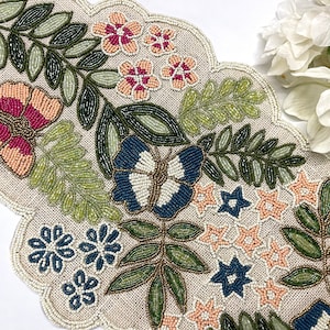 Op de afbeelding: Beaded tafelloper met een geschulpte rand. De loper heeft een bloemen- en vlinderontwerp met roze, blauwe, groene en beige kralen. Het ontwerp omvat bloemen, bladeren en een vlinder.