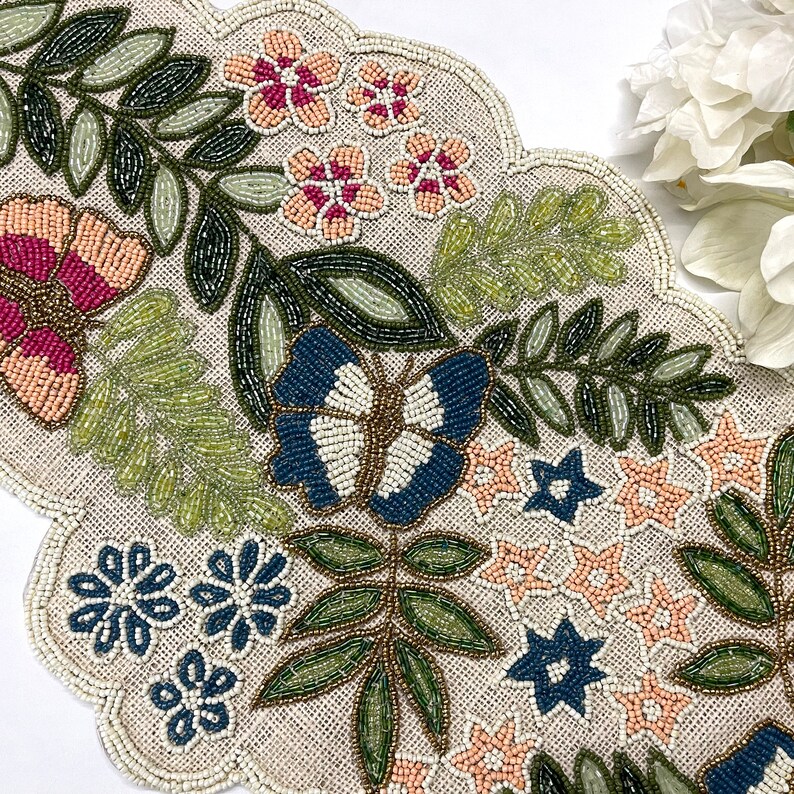 Op de afbeelding: Beaded tafelloper met een geschulpte rand. De loper heeft een bloemen- en vlinderontwerp met roze, blauwe, groene en beige kralen. Het ontwerp omvat bloemen, bladeren en een vlinder.