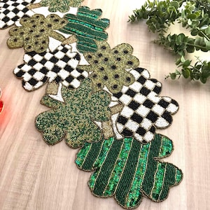 May include: A beaded table runner featuring a clover leaf design. The runner has alternating patterns of green, gold, black and white. Some sections have a checkered pattern, while others have stripes or solid colors. The runner is on a light wood surface.