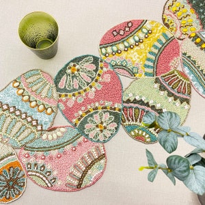 以下が含まれることがあります： カラフルな卵のデザインが施されたビーズ製のテーブルランナー。ランナーはピンク、グリーン、ブルー、イエローの小さなビーズで作られています。卵は湾曲したパターンで配置されており、ランナーの端は卵よりも長く伸びています。