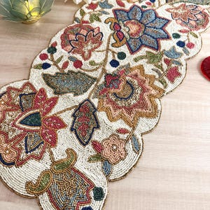 Könnte beinhalten: Cremefarbener Perlen-Tischläufer mit floralem Design. Der Läufer weist komplizierte Muster in Blau-, Rot-, Rosa- und Goldtönen auf. Die Kanten haben ein gewelltes Design. Der Läufer liegt auf einer hellen Holzoberfläche.