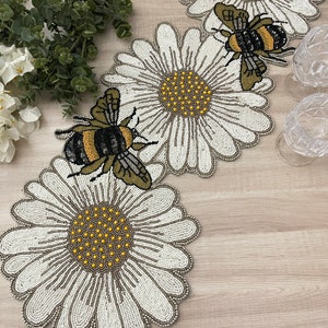 May include: A white beaded table runner with a daisy and bee design. The runner features three large daisies with yellow centers and black and yellow bees.