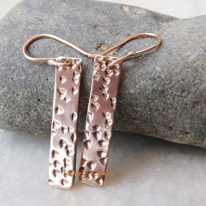 Pode incluir: Um par de brincos de barra martelada em ouro rosa com uma superfície texturizada. Os brincos têm um design simples e minimalista.
