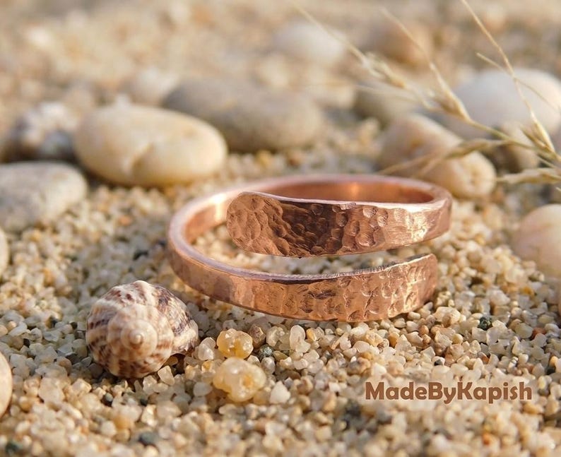 May include: A copper-colored bypass ring featuring a hammered, textured surface. The ring has an open, adjustable design with overlapping ends. The text MadeByKapish is visible in the lower right corner of the image, set against a sandy background.