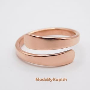 May include: A rose gold-toned bypass ring with a smooth, polished finish and an open, adjustable band design. The metal surface is reflective and uniform in color. The text MadeByKapish is printed in a light brown font below the ring.
