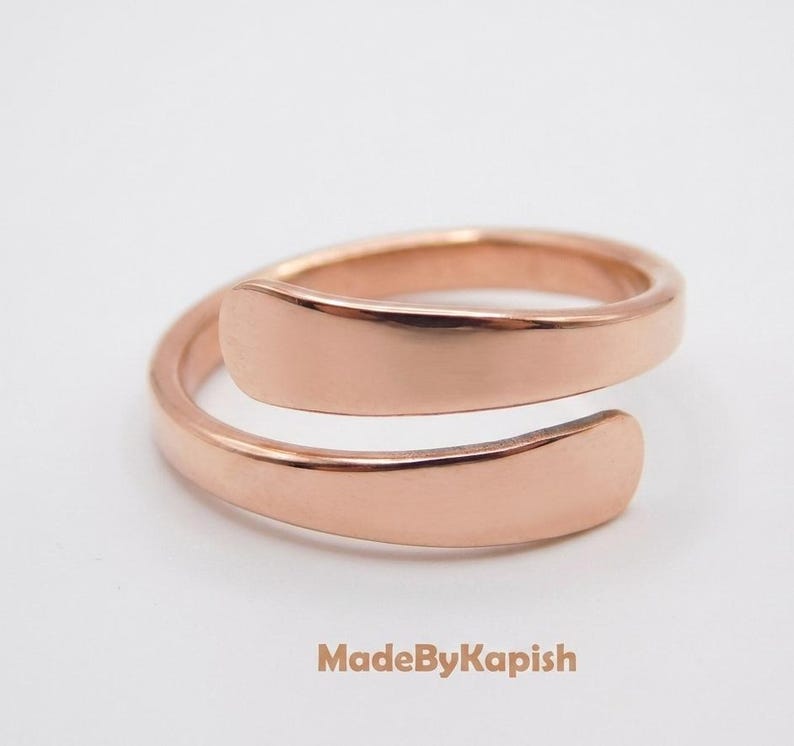May include: A rose gold-toned bypass ring with a smooth, polished finish and an open, adjustable band design. The metal surface is reflective and uniform in color. The text MadeByKapish is printed in a light brown font below the ring.