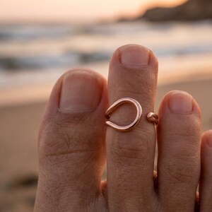 Op de afbeelding: Een open teenring in ros&eacute;goudkleur met een glad, afgerond, druppelvormig ontwerp. De metalen band is gepolijst en reflecterend, met een kleine opening aan de zijkant, en zit om de tweede teen van een voet.