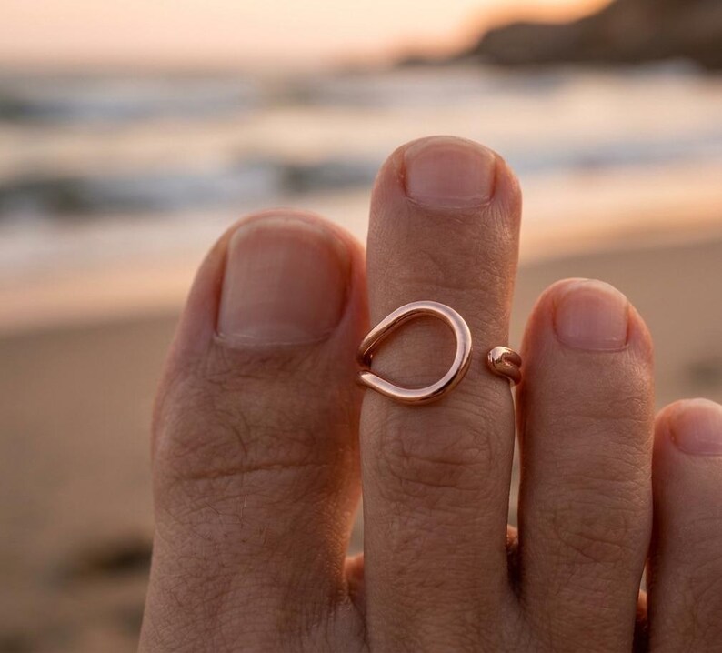 Op de afbeelding: Een open teenring in ros&eacute;goudkleur met een glad, afgerond, druppelvormig ontwerp. De metalen band is gepolijst en reflecterend, met een kleine opening aan de zijkant, en zit om de tweede teen van een voet.