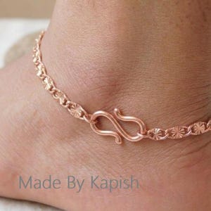 Pode incluir: Uma tornozeleira em tom de ouro rosa com uma corrente delicada de elos circulares texturizados em forma de estrela e um fecho de gancho em forma de S. O metal tem um brilho quente e o design é preso ao tornozelo com o gancho visível.