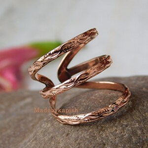 May include: A copper-colored ring with a unique, swirling design. The ring features three bands with an embossed floral pattern. The ring is displayed on a stone surface, with a blurred background of pink and green.