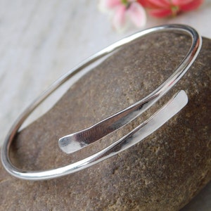 May include: A silver bangle bracelet with a simple, minimalist design. The bracelet is made of a single, continuous piece of metal that is bent into a circle. The ends of the bracelet are joined together with a small, subtle overlap.