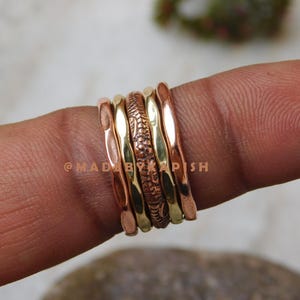 May include: A stack of four rings made of copper and brass. The rings are hammered and have a textured surface. The rings are worn on a finger.