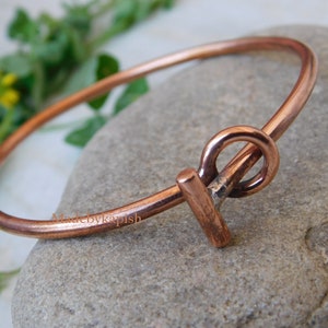 May include: A simple copper bangle bracelet with a unique, handmade clasp. The clasp is a twisted copper wire that forms a circle and a bar.