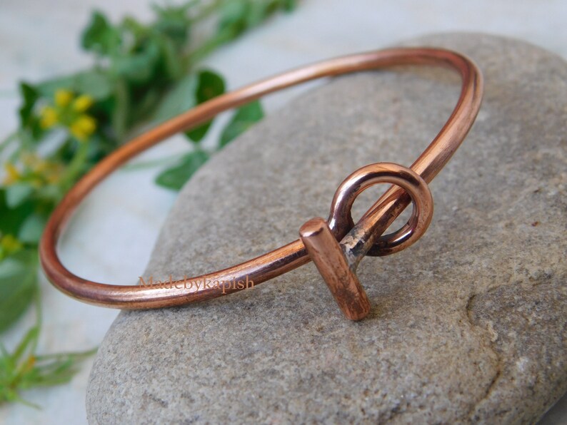 May include: A simple copper bangle bracelet with a unique, handmade clasp. The clasp is a twisted copper wire that forms a circle and a bar.