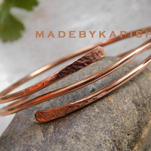 May include: A copper-colored arm cuff bracelet with a hammered texture on the ends. The bracelet wraps around the arm in a spiral design. The text "MADEBYKAPISH" is visible in the image.