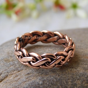 Op de afbeelding: Een gevlochten koperen ring met een gladde, gepolijste afwerking. De ring is gemaakt van een enkel stuk koperdraad dat is geweven tot een uniek en ingewikkeld ontwerp.