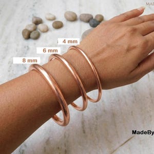 Puede incluir: Tres brazaletes de cobre pulido y liso en la muñeca, con grosores distintos etiquetados como 4 mm, 6 mm y 8 mm. Las pulseras metálicas tienen un perfil redondeado y un tono marrón rojizo cálido, mostrando claramente las diferentes anchuras.