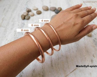 May include: Three smooth, polished copper bangles worn on the wrist, featuring distinct thicknesses labeled as 4 mm, 6 mm, and 8 mm. The metallic bracelets have a rounded profile and a warm, reddish-brown tone, showcasing the varying widths clearly.