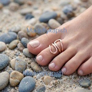May include: A rose gold-toned toe ring featuring a double-loop, open-ended design with smooth, rounded edges. The ring is worn on the second toe, showcasing a metallic finish that contrasts with the natural skin tone and the surrounding pebbles.