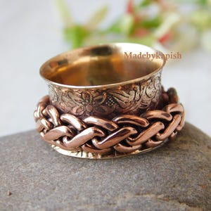 Könnte beinhalten: Ein drehbarer Ring aus Gold und Kupfer mit einem filigranen Blumendesign. Der Ring besteht aus zwei Bändern, eines mit einem geflochtenen Muster und das andere mit einem Blumendesign. Der Ring liegt auf einem grauen Stein.