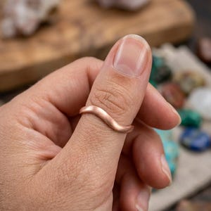 May include: A copper-colored ring with a wavy design. The ring is worn on a finger, showcasing its smooth, curved lines. The ring's warm hue contrasts with the skin tone, highlighting its unique shape. The ring is a piece of jewelry.