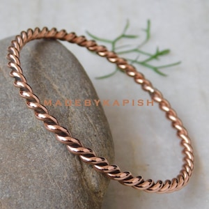 May include: A simple copper bangle bracelet with a twisted design. The bracelet is made of a single piece of copper wire that has been twisted together to create a rope-like effect.