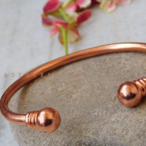 Puede incluir: Un brazalete de cobre con extremos redondeados. El brazalete está hecho de una sola pieza de cobre y tiene un acabado liso y pulido.