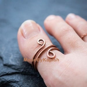 May include: A copper wire ring featuring an open, bypass-style design with two coiled ends. The metal has a warm, reddish-brown tone and a smooth, rounded finish. The text MadeByKapish is visible across the centre of the ring band.