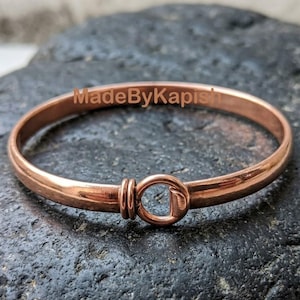 May include: A polished copper bangle bracelet featuring a circular hook-and-eye style clasp mechanism. The metal band has a smooth, reflective surface and is marked with the text MadeByKapish in a light brown font across the center of the piece.