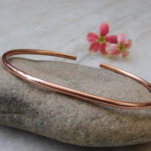 Puede incluir: Un brazalete simple de cobre pulido. El brazalete es delgado y tiene un acabado liso y redondeado.